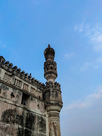 A historical stone tower with intricate carvings on its facade stands against a bright blue sky. The architecture displays a blend of artistic details with repeating geometric patterns and ornate designs. Weathered surfaces contribute to an ancient and timeless appeal.