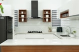 A modern kitchen with a sleek design, featuring white countertops and cabinetry. In the center is a black stovetop with a hood above. Wooden shelves contain small storage jars labeled with various contents. A black refrigerator is on one side, while small potted plants and kitchen appliances are on the counter.