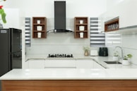 A modern kitchen with a sleek design, featuring white countertops and cabinetry. In the center is a black stovetop with a hood above. Wooden shelves contain small storage jars labeled with various contents. A black refrigerator is on one side, while small potted plants and kitchen appliances are on the counter.