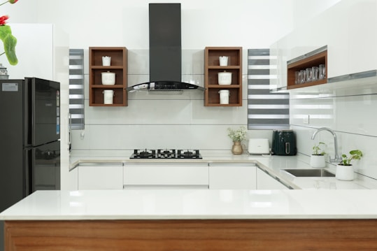 A modern kitchen with a sleek design, featuring white countertops and cabinetry. In the center is a black stovetop with a hood above. Wooden shelves contain small storage jars labeled with various contents. A black refrigerator is on one side, while small potted plants and kitchen appliances are on the counter.