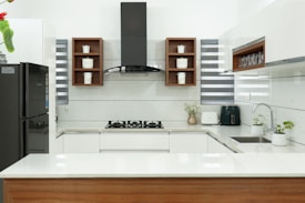 A modern kitchen with a sleek design, featuring white countertops and cabinetry. In the center is a black stovetop with a hood above. Wooden shelves contain small storage jars labeled with various contents. A black refrigerator is on one side, while small potted plants and kitchen appliances are on the counter.