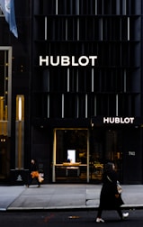 A storefront of a high-end watch brand featuring the name Hublot in bold letters on a sleek, dark facade. Two people are walking on the sidewalk, with one carrying an orange shopping bag and the other a beige handbag. The entrance to the store is illuminated, showcasing a modern and luxurious interior.