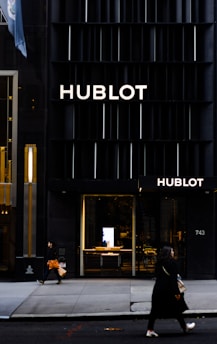 A storefront of a high-end watch brand featuring the name Hublot in bold letters on a sleek, dark facade. Two people are walking on the sidewalk, with one carrying an orange shopping bag and the other a beige handbag. The entrance to the store is illuminated, showcasing a modern and luxurious interior.