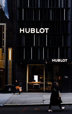 A storefront of a high-end watch brand featuring the name Hublot in bold letters on a sleek, dark facade. Two people are walking on the sidewalk, with one carrying an orange shopping bag and the other a beige handbag. The entrance to the store is illuminated, showcasing a modern and luxurious interior.