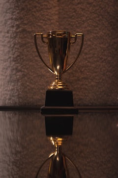 A shiny golden trophy with two handles resting on a black base is prominently displayed. The surface beneath provides a reflective view, adding depth to the composition. The background is textured and softly lit, offering a contrast to the gleaming trophy.