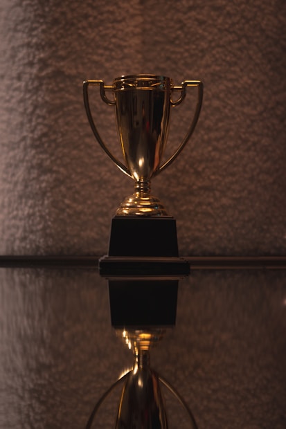 A shiny golden trophy with two handles resting on a black base is prominently displayed. The surface beneath provides a reflective view, adding depth to the composition. The background is textured and softly lit, offering a contrast to the gleaming trophy.