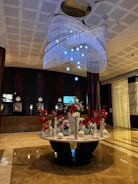 The image features a spacious and elegant hotel lobby with a polished marble floor. There is a large, ornate chandelier hanging from the ceiling, adorned with multiple layers of crystal-like beads and blue lights. Below the chandelier stands a round marble table that holds an assortment of flower vases filled with red and white flowers. The background displays a dark-colored reception area with several clocks, screens, and lamps on the counter.