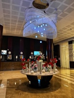 The image features a spacious and elegant hotel lobby with a polished marble floor. There is a large, ornate chandelier hanging from the ceiling, adorned with multiple layers of crystal-like beads and blue lights. Below the chandelier stands a round marble table that holds an assortment of flower vases filled with red and white flowers. The background displays a dark-colored reception area with several clocks, screens, and lamps on the counter.