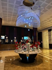 The image features a spacious and elegant hotel lobby with a polished marble floor. There is a large, ornate chandelier hanging from the ceiling, adorned with multiple layers of crystal-like beads and blue lights. Below the chandelier stands a round marble table that holds an assortment of flower vases filled with red and white flowers. The background displays a dark-colored reception area with several clocks, screens, and lamps on the counter.