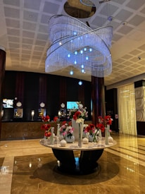 The image features a spacious and elegant hotel lobby with a polished marble floor. There is a large, ornate chandelier hanging from the ceiling, adorned with multiple layers of crystal-like beads and blue lights. Below the chandelier stands a round marble table that holds an assortment of flower vases filled with red and white flowers. The background displays a dark-colored reception area with several clocks, screens, and lamps on the counter.