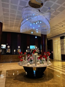 The image features a spacious and elegant hotel lobby with a polished marble floor. There is a large, ornate chandelier hanging from the ceiling, adorned with multiple layers of crystal-like beads and blue lights. Below the chandelier stands a round marble table that holds an assortment of flower vases filled with red and white flowers. The background displays a dark-colored reception area with several clocks, screens, and lamps on the counter.