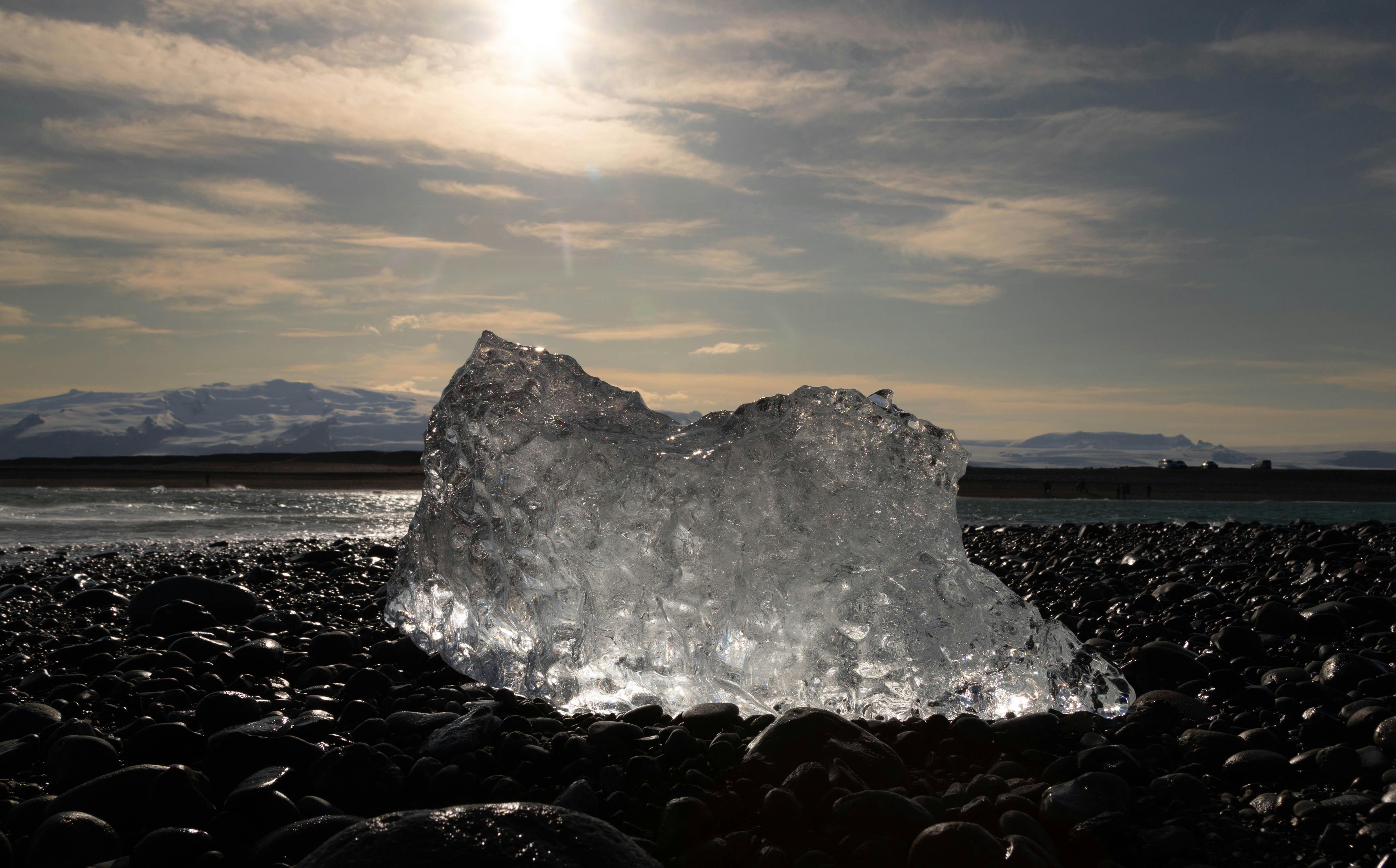 a large iceberg sitting on top of a rocky beach, A striking piece of glacial ice sits prominently on a black pebble beach, its crystalline structure sparkling under the sunlight. The contrast between the ice