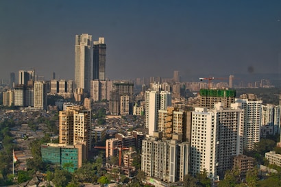 A cityscape featuring numerous high-rise buildings under a clear sky. The image shows densely packed skyscrapers and residential towers, with construction cranes visible atop some structures, indicating ongoing development. Lush greenery intersperses the urban environment, adding contrast to the predominantly concrete landscape.