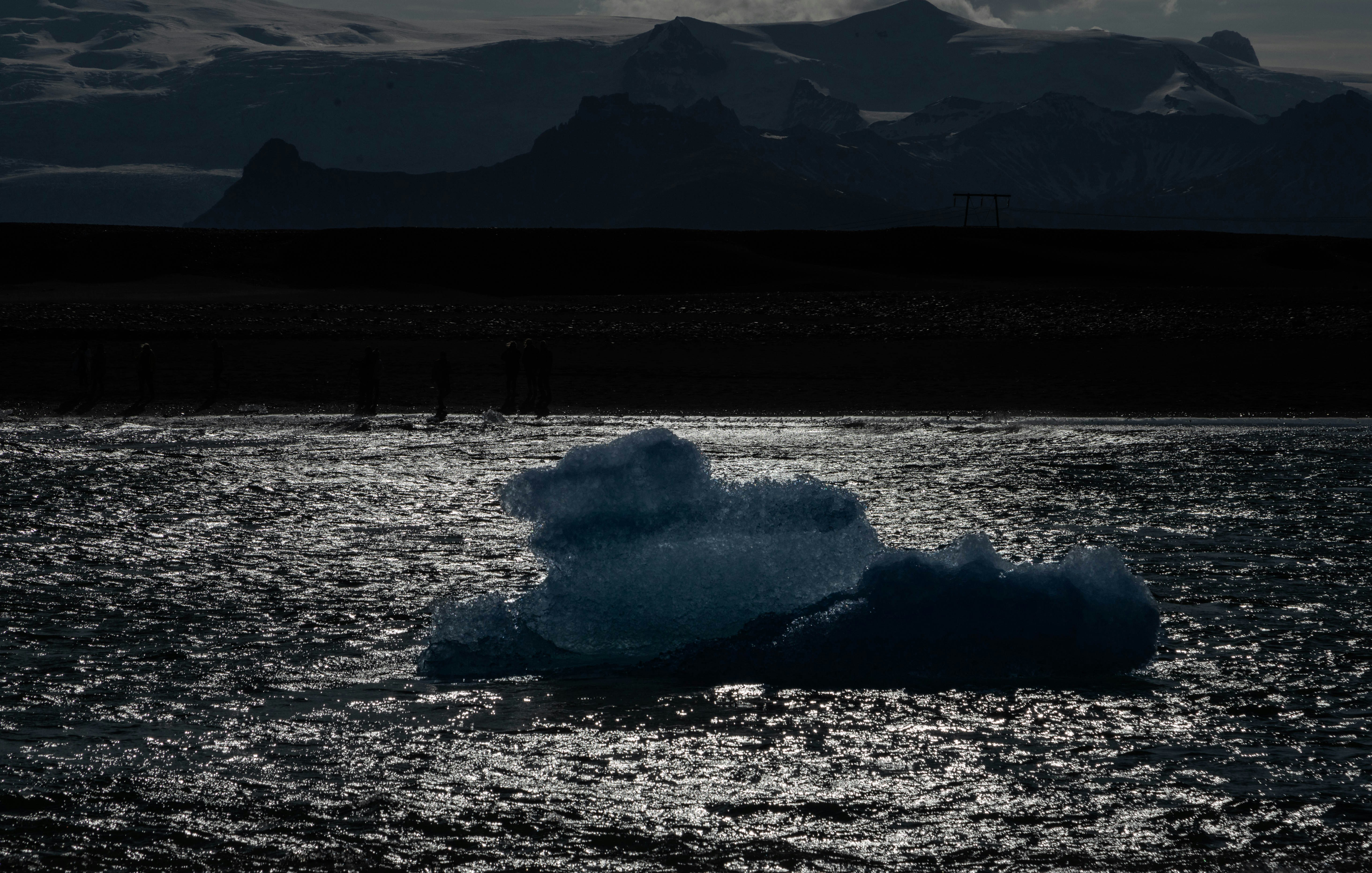 a large iceberg floating in the middle of a lake, A solitary iceberg floats on a glimmering body of water, silhouetted against the sparkling light. In the distance, figures are dwarfed by the expansive landscape, with majestic mountains looming under a soft-lit sky, highlighting the stark contrasts of the Arctic wilderness.