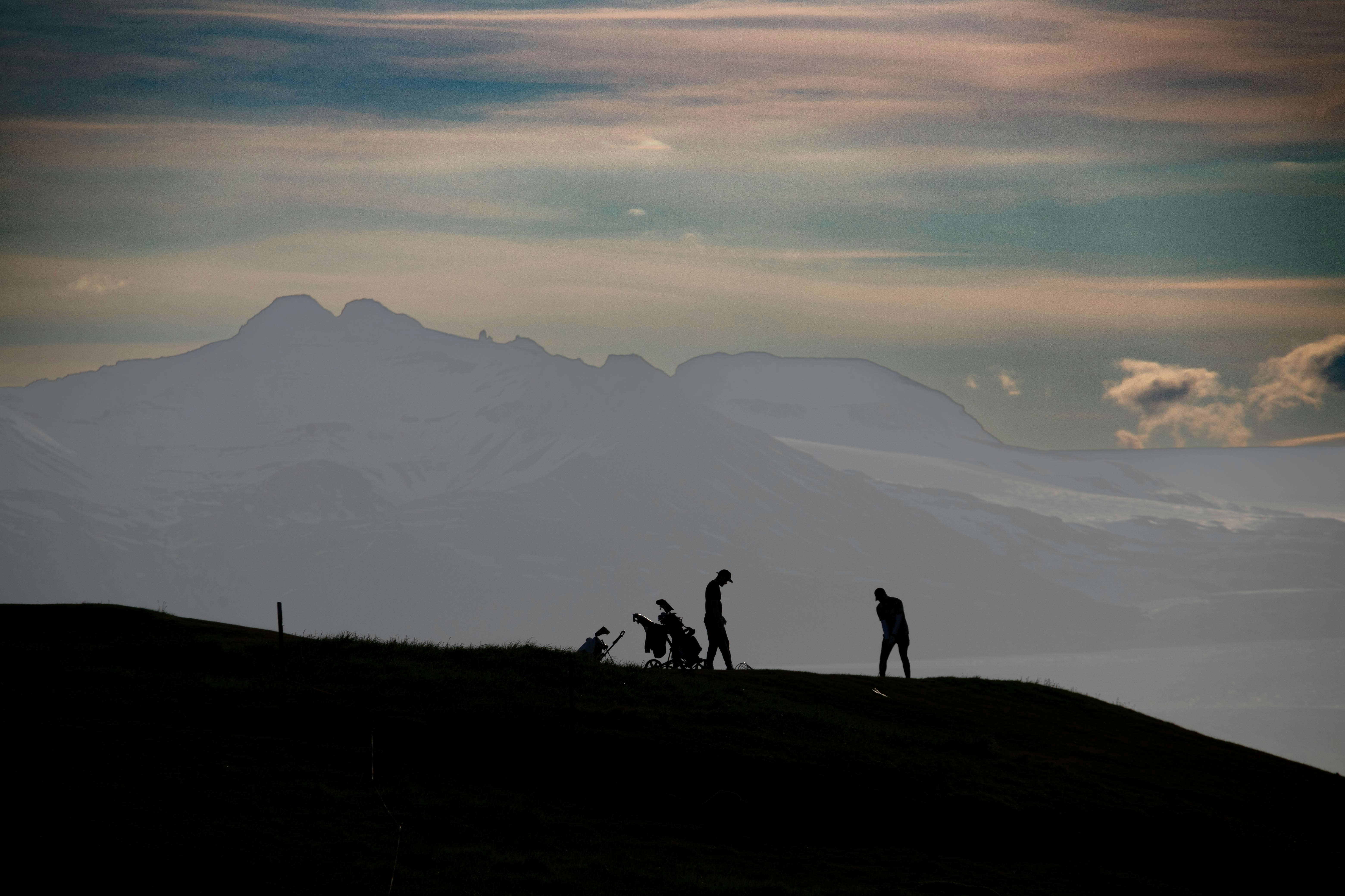 a group of people standing on top of a hill, Silhouettes of three people, one pushing a stroller, on a hill with a dramatic mountain range in the background under a dusky sky with soft clouds.