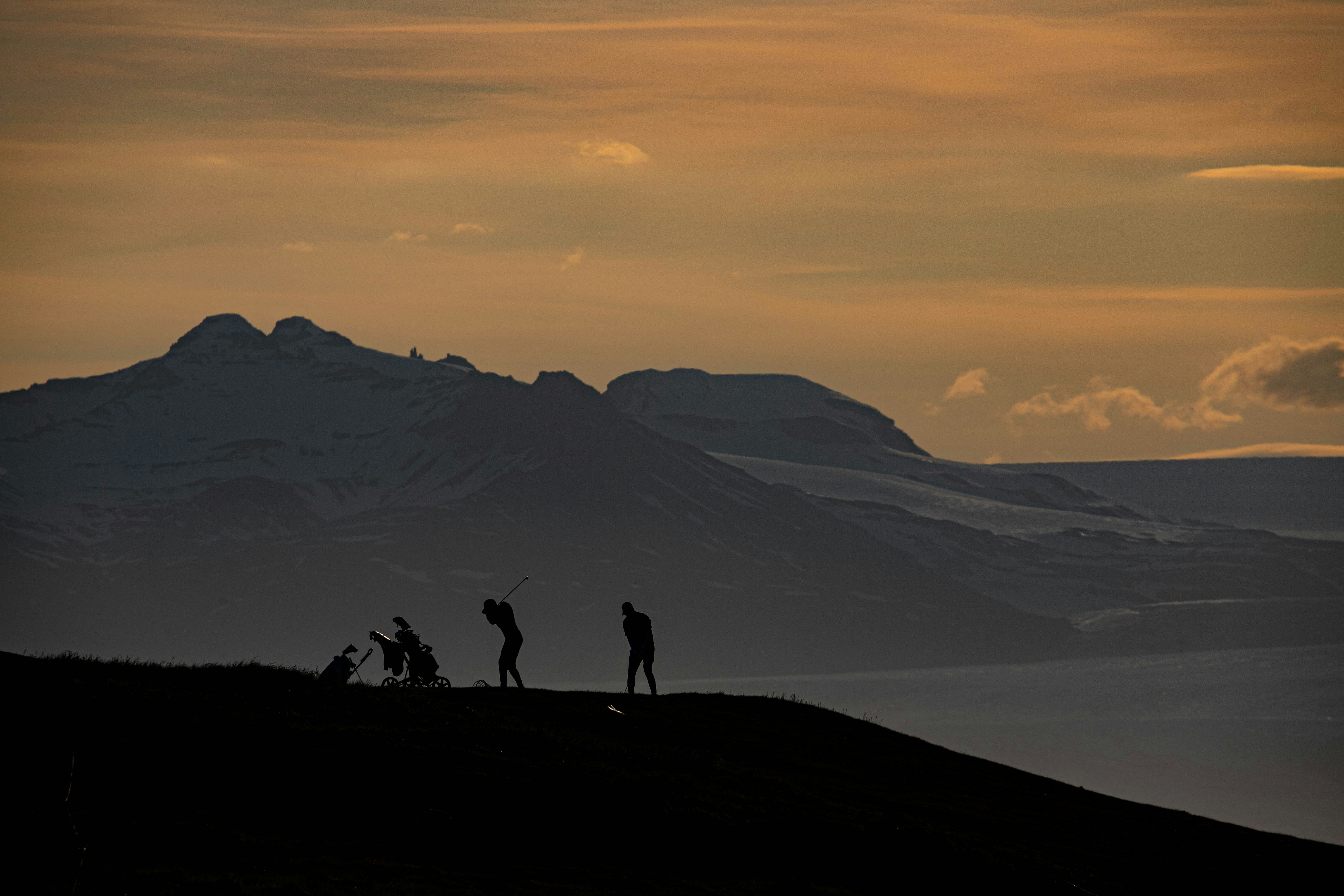 Silhouettes of three golfers and two golf bags on a hill, with a striking mountain range backdrop under a dusky sky.