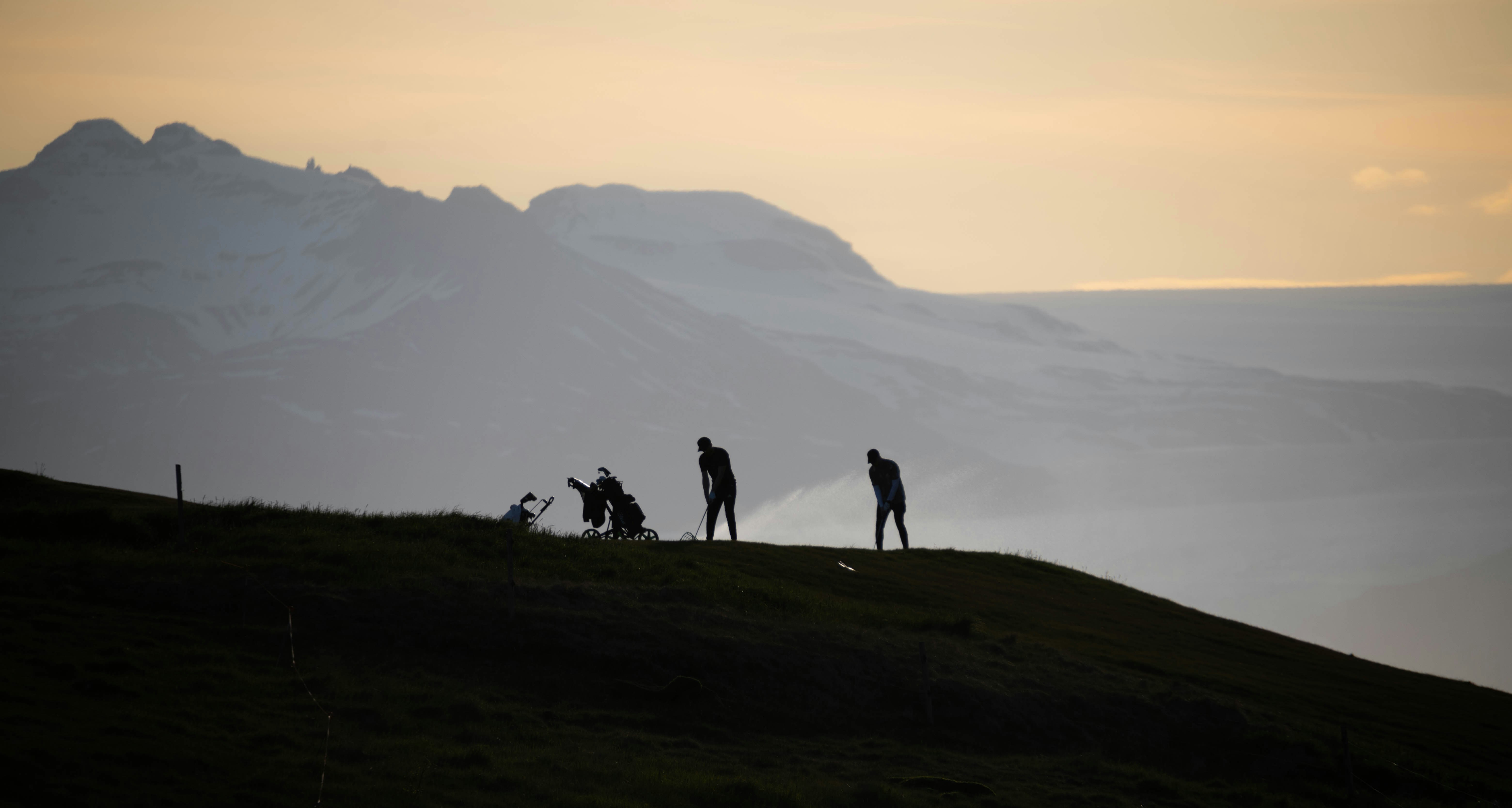 Golden sunset hues bathe a mountainous landscape where silhouetted figures appear with golf equipment on a gently sloping hill.