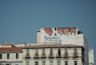 A large building with graffiti art on its upper section, including bold red-and-black lettering. The rest of the building displays a more subdued beige tone, with multiple balconies and windows. The sky is clear and blue, forming a stark backdrop against the urban scene.