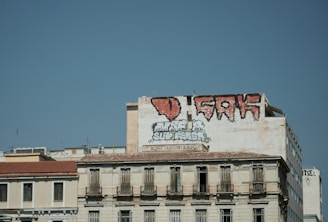 A large building with graffiti art on its upper section, including bold red-and-black lettering. The rest of the building displays a more subdued beige tone, with multiple balconies and windows. The sky is clear and blue, forming a stark backdrop against the urban scene.