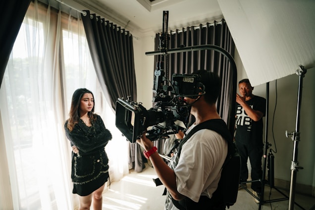 A scene captures a film production setup with a young woman standing beside large curtains, her arms crossed and looking thoughtful. A cameraman in the foreground operates a professional camera on a rig, while another crew member stands in the background near lighting equipment.