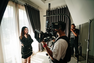 A scene captures a film production setup with a young woman standing beside large curtains, her arms crossed and looking thoughtful. A cameraman in the foreground operates a professional camera on a rig, while another crew member stands in the background near lighting equipment.