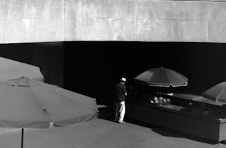 A solitary figure stands under a large umbrella, positioned in front of a dark stall with what appears to be a selection of items on display. The scene is captured in monochrome, with high contrast between the shaded and exposed areas, setting a mood of solitude or contemplation.