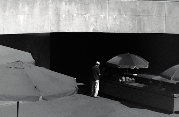 A solitary figure stands under a large umbrella, positioned in front of a dark stall with what appears to be a selection of items on display. The scene is captured in monochrome, with high contrast between the shaded and exposed areas, setting a mood of solitude or contemplation.