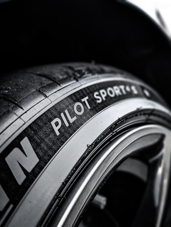 A close-up view of a car tire with the text 'Pilot Sport' clearly visible on the sidewall. The tire texture and tread details are prominent, set against a dark background that showcases the metallic sheen of the rim.