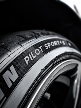 A close-up view of a car tire with the text 'Pilot Sport' clearly visible on the sidewall. The tire texture and tread details are prominent, set against a dark background that showcases the metallic sheen of the rim.