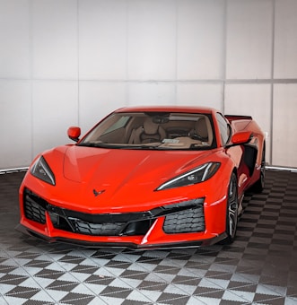 A vibrant red sports car with a sleek design and aerodynamic features is showcased indoors. The car's aggressive front styling includes sharp headlights and a prominent grille, and it is set against a checkered floor.