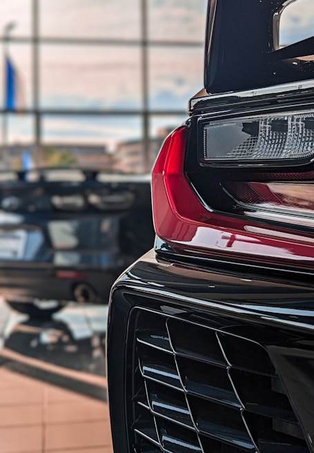Close-up view of a car's rear light assembly and bumper, with another car and large windows in the background, creating a showroom setting.