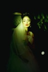 A woman with red lips wearing a wedding dress and veil is softly illuminated against a dark background. Her expression is serene, and she is positioned in a way that highlights her face and the fine details of her dress. The lighting creates a dramatic effect, casting shadows and adding depth to the scene.