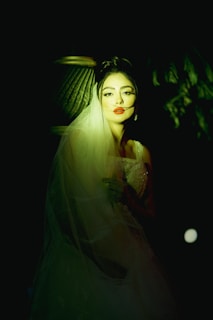 A woman with red lips wearing a wedding dress and veil is softly illuminated against a dark background. Her expression is serene, and she is positioned in a way that highlights her face and the fine details of her dress. The lighting creates a dramatic effect, casting shadows and adding depth to the scene.