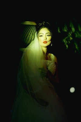 A woman with red lips wearing a wedding dress and veil is softly illuminated against a dark background. Her expression is serene, and she is positioned in a way that highlights her face and the fine details of her dress. The lighting creates a dramatic effect, casting shadows and adding depth to the scene.
