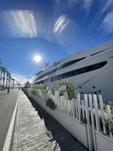 A large luxury yacht is docked along a waterfront with palm trees on the left side and a bright sun in a clear blue sky. There is a paved sidewalk next to a white fence with small shrubs, and the shadows stretch long in front due to the sun's low angle.