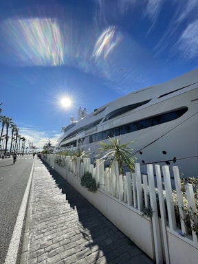 A large luxury yacht is docked along a waterfront with palm trees on the left side and a bright sun in a clear blue sky. There is a paved sidewalk next to a white fence with small shrubs, and the shadows stretch long in front due to the sun's low angle.