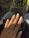 A hand with long, almond-shaped nails painted in a light, creamy beige color that features small decorative gems on each nail. The hand rests on what appears to be a black leather surface, with a background of denim and polka-dot patterns in soft focus.