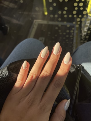 A hand with long, almond-shaped nails painted in a light, creamy beige color that features small decorative gems on each nail. The hand rests on what appears to be a black leather surface, with a background of denim and polka-dot patterns in soft focus.