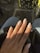 A hand with long, almond-shaped nails painted in a light, creamy beige color that features small decorative gems on each nail. The hand rests on what appears to be a black leather surface, with a background of denim and polka-dot patterns in soft focus.