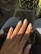 A hand with long, almond-shaped nails painted in a light, creamy beige color that features small decorative gems on each nail. The hand rests on what appears to be a black leather surface, with a background of denim and polka-dot patterns in soft focus.