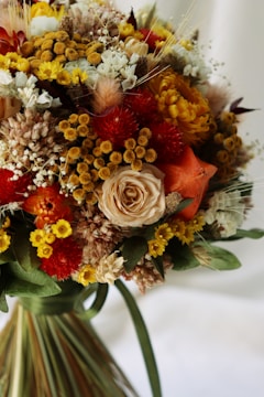 A bouquet of dried and fresh flowers featuring a variety of textures and colors. It includes orange and red blooms, yellow pom-pom-like flowers, a central beige rose, and vibrant green leaves.