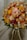 A vibrant bouquet of various flowers featuring shades of orange, yellow, pink, and red. The bouquet includes roses, chrysanthemums, and dried flowers, along with green leaves. The composition is dense and artistically arranged, displaying a textured and colorful appearance.