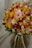 A vibrant bouquet of various flowers featuring shades of orange, yellow, pink, and red. The bouquet includes roses, chrysanthemums, and dried flowers, along with green leaves. The composition is dense and artistically arranged, displaying a textured and colorful appearance.
