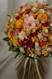 A vibrant bouquet of various flowers featuring shades of orange, yellow, pink, and red. The bouquet includes roses, chrysanthemums, and dried flowers, along with green leaves. The composition is dense and artistically arranged, displaying a textured and colorful appearance.