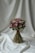 A bouquet of flowers is elegantly arranged with a mix of pink, purple, and green hues. The bouquet is tied with a maroon ribbon and is set against a softly draped white fabric backdrop, creating a serene and sophisticated presentation.