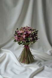 A bouquet of flowers is elegantly arranged with a mix of pink, purple, and green hues. The bouquet is tied with a maroon ribbon and is set against a softly draped white fabric backdrop, creating a serene and sophisticated presentation.