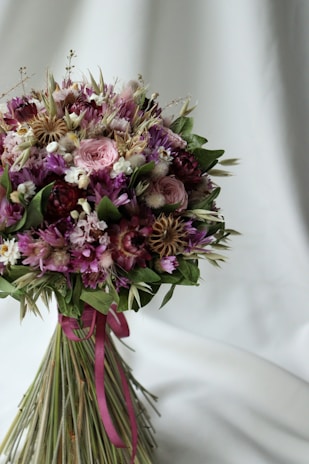 A bouquet of flowers with a mixture of pink, purple, and white blooms, featuring various types such as roses and daisies. The bouquet is wrapped with a purple ribbon and has a green foliage background against a light, soft draped fabric.