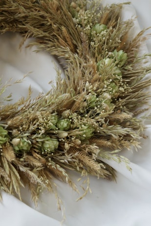 A close-up view of a wreath made from dried grasses and greenish buds arranged in a circular pattern. The wreath lies on a soft, white fabric background, creating a contrast with its natural, earthy tones.