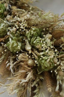 A detailed arrangement of dried wheat stalks and green flowers, interspersed with small white buds. The textures and colors of the plants create a rustic and natural aesthetic.