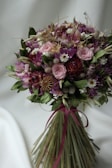 A bouquet of dried flowers featuring a variety of textures and colors, including soft pink roses, vibrant purple blooms, small white flowers, and green leaves. The stems are elegantly tied with a dark pink ribbon.